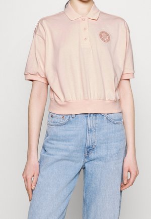 Persoon draagt een lichtroze cropped poloshirt met knopen en een klein rond logo, gecombineerd met high-waisted lichtblauwe spijkerbroek.