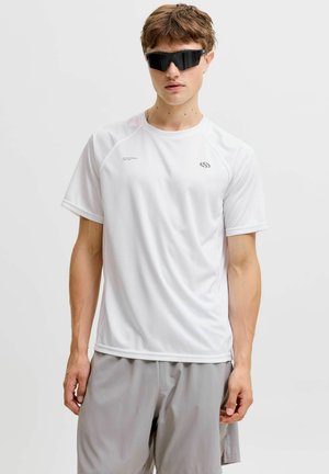 Jeune homme portant des lunettes de soleil noires enveloppantes, une chemise blanche à manches courtes et un short gris clair sur fond uni.