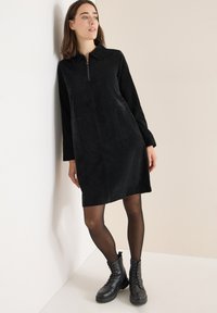 Vestito in corduroy nero con chiusura a zip, maniche lunghe, due tasche frontali e colletto, abbinato a stivali neri con lacci.