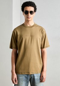 Barna, rövid ujjú póló kerek nyakkal, rajta a "SCOTCH & SODA" logó, világoskék farmernadrággal párosítva. A modellen fekete napszemüveg van.