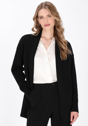 Schwarze offenfallende Strickjacke aus weichem Material, mit langen Ärmeln und lockerer Passform, kombiniert mit einer weißen Bluse und schwarzen Hosen.