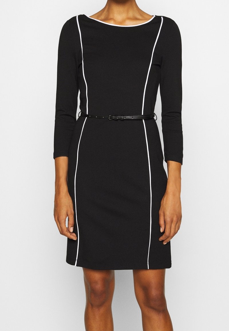 Robe noire à manches longues, avec des accents de passepoil blanc sur les côtés et une fine ceinture noire. Fabriquée dans un tissu lisse et structuré.