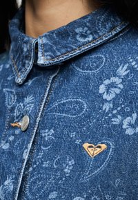 Tuvuņskats uz džinsa krekla apkakli ar ziedu un paisley rakstiem, sudraba pogu un mazu bēšas krāsas sirdsdilu loģotipa izšuvumu.