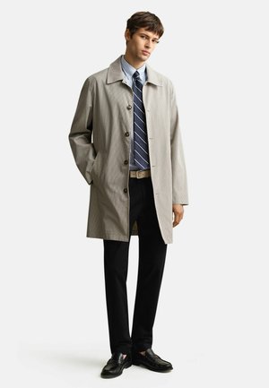 Homme debout portant un pardessus à carreaux beige, une chemise habillée bleue, une cravate rayée, un pantalon noir et des mocassins noirs sur un fond uni.