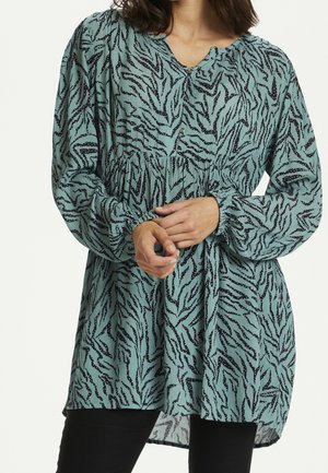 Kobieta ubrana w luźną, turkusową bluzkę z długim rękawem, z czarnym abstrakcyjnym wzorem zebry, guzikami i marszczonym stanem, dłonie splecione na przodzie.