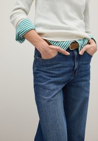 Weißer Pullover mit gestreiftem grünen Bündchen, über einem blau-weiß gestreiften Hemd getragen, kombiniert mit dunkelblauen Jeans und einem braunen Gürtel.