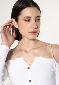 Top bianco a costine con scollo a V, spalline in catena argentata e dettagli di bottoni in evidenza. Il tessuto sembra morbido con una finitura texturizzata.