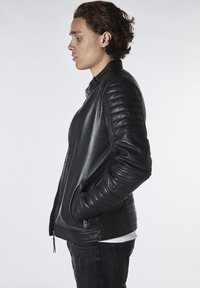 Chaqueta de cuero negra con hombros acolchados y bolsillos laterales. Presenta un cierre de cremallera y una textura suave. Lleva puesta sobre una camisa blanca.