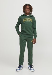 Sudadera verde con pantalones de chándal a juego y texto amarillo "JACK & JONES". El material es suave y el ajuste es casual. Las zapatillas tienen detalles en azul, blanco y negro.