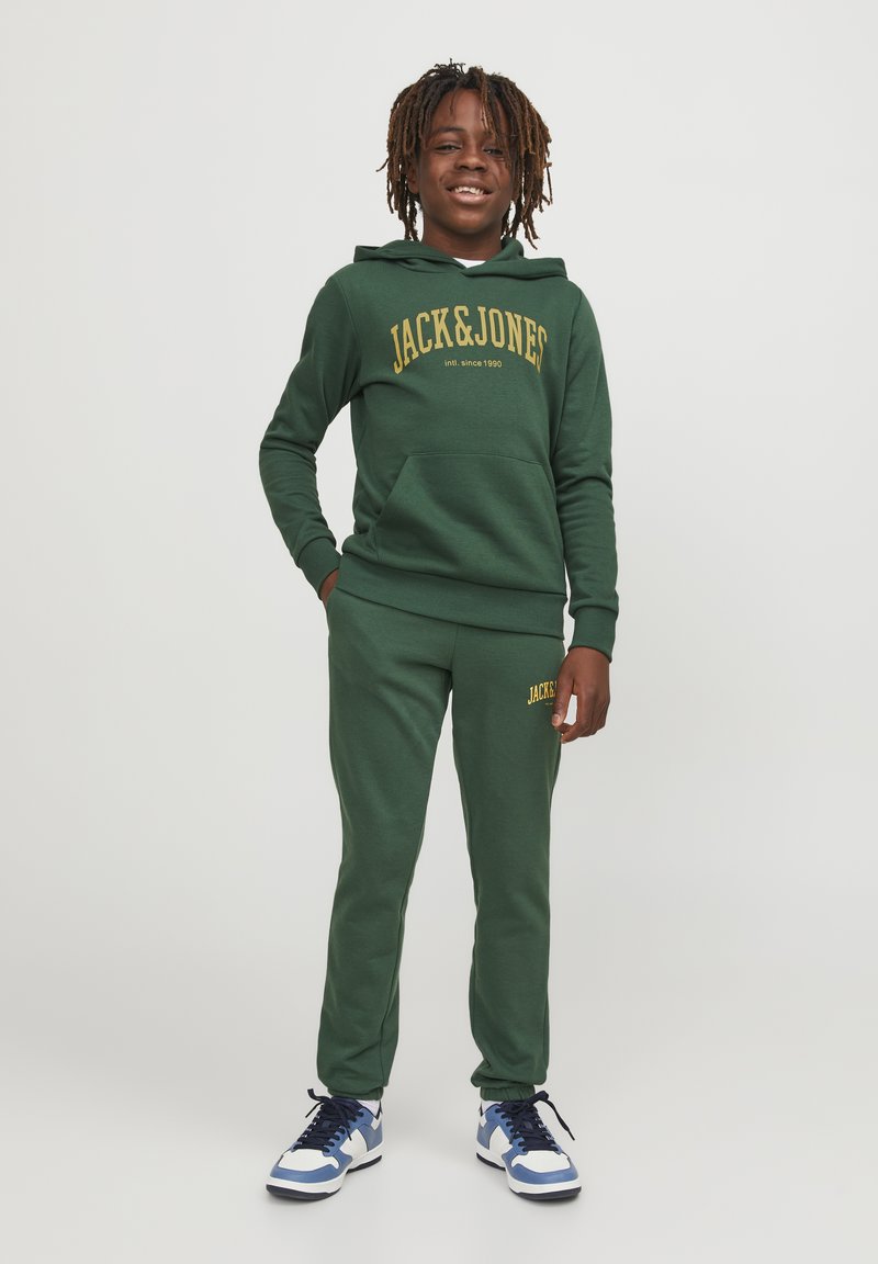 Sudadera verde con pantalones de chándal a juego y texto amarillo "JACK & JONES". El material es suave y el ajuste es casual. Las zapatillas tienen detalles en azul, blanco y negro.