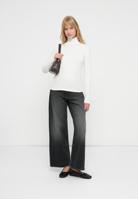 Bela turtleneck majica z dolgimi rokavi, v kombinaciji s širokimi temno sivimi kavbojkami in črnimi balerinkami. Drži majhno rjavo torbico.