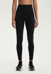 Magas derekú fekete leggings sima textúrával, fehér oldalsávokkal és alakformáló dizájnnal, fekete sportcipővel párosítva.