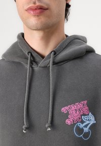 Sweatshirt cinza com capuz e cordões, apresentando um logótipo bordado em rosa e azul e um gráfico de cerejas no peito. Tecido macio e texturizado.