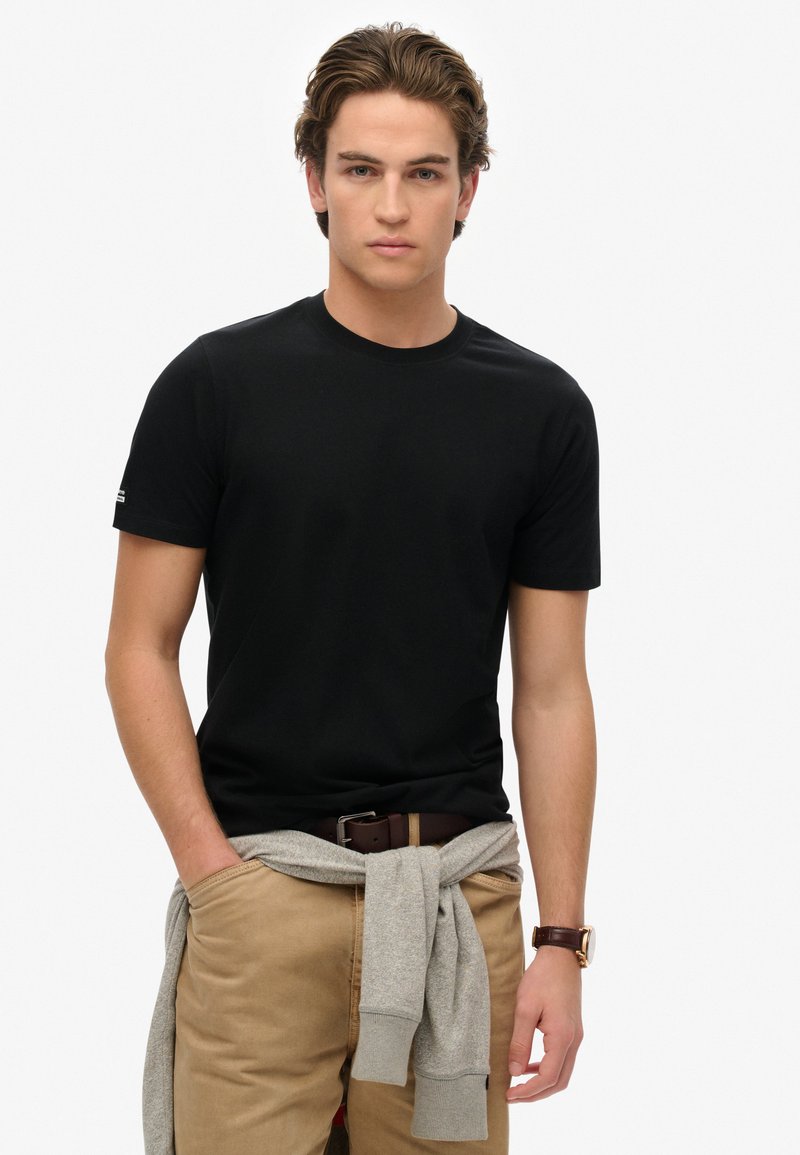 Sort T-shirt med korte ærmer og rund hals, lavet af glat bomuld. En grå sweatshirt er bundet omkring livet. Brun bælte og ur er synligt.