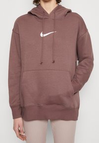 Person klädd i en brun Nike-hoodie med vit logotyp, frampocket och dragsnören, tillsammans med ljusa ribbade byxor.
