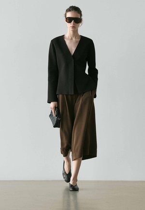 Kvinde iført sort blazer, store solbriller, brune vide bukser afkortet ved anklerne, sorte flade sko og holder en lille sort clutch mod en grå baggrund.