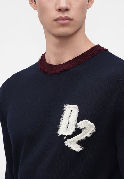 Jeune homme portant un pull bleu marine avec un col rond bordeaux effiloché et le numéro 02 blanc effiloché sur la poitrine.