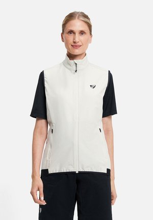 Frau mit hellem Haar, die eine weiße, ärmellose Weste mit Reißverschluss über schwarzen Kurzarmshirt trägt und vor einem schlichten weißen Hintergrund steht.