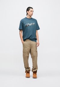 Camisa de manga corta color azul verdoso con el logo metálico "Carhartt", combinada con pantalones cargo color beige y botas de trabajo marrones. Diseño sencillo y de corte relajado.