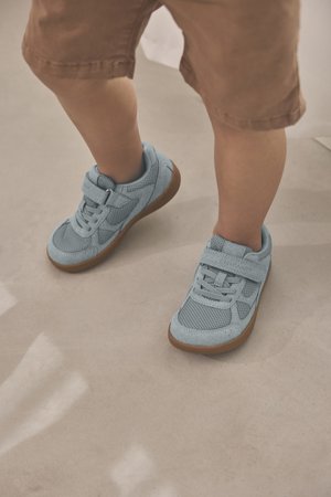 Sneakers azzurro chiaro in mesh e suede, con cinturino in velcro e suola in gomma con dettagli marroni. Indossate con pantaloncini beige.