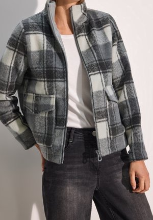 Kobieta w szarej i białej kratowanej polarowej kurtce zapinanej na zamek, na białej koszulce i z czarnymi, wypranymi dżinsami, stoi z jedną ręką na biodrze.