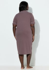 Mujer de pie descalza de espaldas, vistiendo un vestido color malva hasta la rodilla con manga corta y ribetes blancos en el cuello y las mangas.