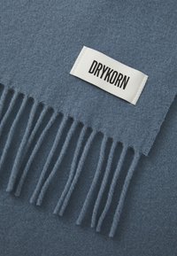Plava vuna šal s resicama na rubovima. Oznaka s natpisom "DRYKORN" u debelim crnim slovima na bijeloj pozadini. Mekana tekstura s ravnomjernom završnom obradom.