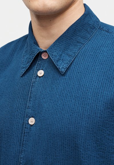 Κοντινό πλάνο ενός ατόμου που φοράει μια μπλε πουκάμισο με υφή και γιακά με τρία κουμπιά, ένα κουμπί είναι καφέ και τα άλλα δύο μπεζ.