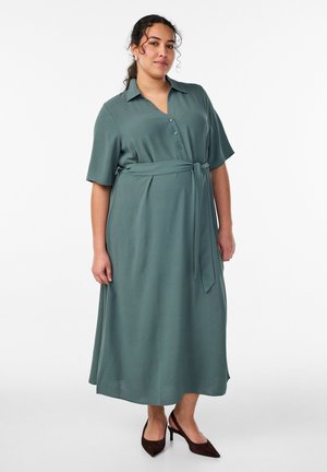 Vrouw met krullend haar, een groene midi-jurk met korte mouwen en ceintuur, en donkere slingback hakschoenen, staand tegen een witte achtergrond.