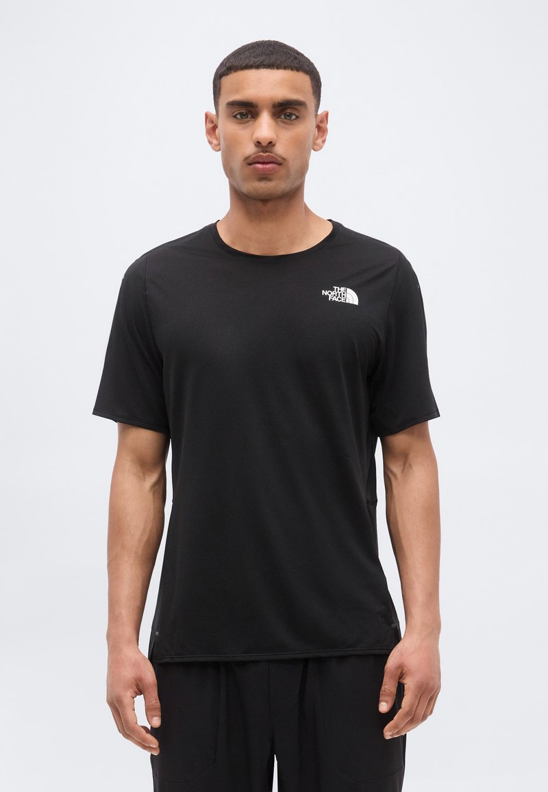 Άνδρας που φοράει μαύρο μονόχρωμο t-shirt The North Face στέκεται μπροστά σε ανοιχτό γκρι φόντο, με ουδέτερη έκφραση.