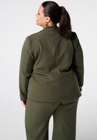 Olijfgroene pak met een op maat gemaakt blazer en wijde broek. De blazer heeft een geknipt kraag en een gladde stof met een lichte glans.