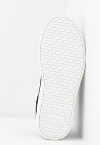 Semelle blanche texturée d'une sneaker Levi's avec logo visible, chaussure pressée contre un mur blanc, vue de dessous.