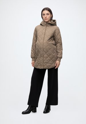 Femme portant un manteau matelassé marron avec capuche et poches, un pantalon noir à jambes larges et des bottines noires, debout devant un fond uni.