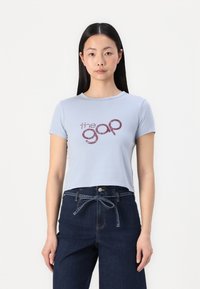 Camiseta corta de color azul claro con mangas cortas y cuello redondo, con el texto "the gap" en rojo oscuro, combinada con pantalones de mezclilla oscuros.