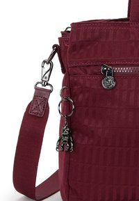 Sac bordeaux avec une finition texturée, comportant une poche zippée, des accessoires en argent et un charm singe noir attaché à la sangle.
