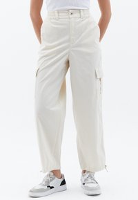 Jasno beżowe spodnie cargo o luźnym kroju, z bocznymi kieszeniami, wysokim stanem i fakturowanym materiałem. Noszone z białymi trampkami.