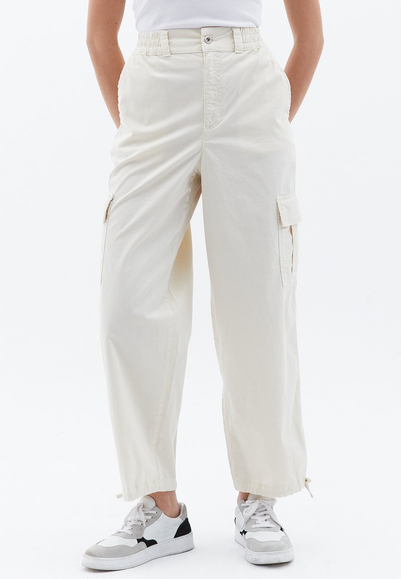 Jasno beżowe spodnie cargo o luźnym kroju, z bocznymi kieszeniami, wysokim stanem i fakturowanym materiałem. Noszone z białymi trampkami.