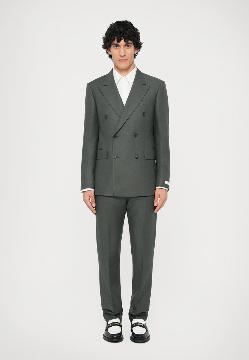 Divrindu tums zaļš uzvalks ar pielāgotu piegriezumu, ar izgrieztām atloks un diviem priekšējiem kabatām. Pāris melnu un baltu kurpju ar zelta akcentiem.