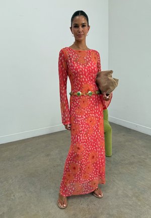 Γυναίκα που στέκεται μέσα σε κλειστό χώρο, φορώντας μακρύ κόκκινο φόρεμα με πορτοκαλί λουλουδάτα σχέδια και πράσινη ζώνη, κρατώντας μια μπεζ τσαντούλα χειρός.