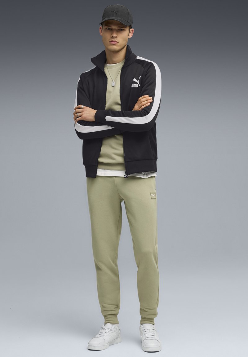 Sort zip-jakke med hvide striber, lys grøn T-shirt, matchende bukser og hvide sneakers. Har Puma-branding. Glat tekstur.