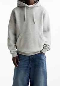 Sweat à capuche gris clair avec une poche frontale, des cordons de serrage et des poignets côtelés, associé à un jean bleu ample. Texture en mélange de coton.