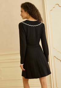 Fekete ruha bordázott, hosszú ujjakkal, pliszírozott szoknyarésszel és hullámos, fehér szegéllyel a nyakkivágásnál. Simas textúrájú, deréknál testhez simuló.
