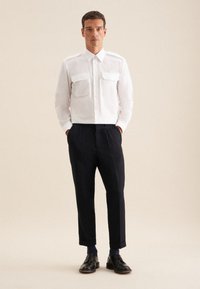Witte button-up blouse met twee borstzakken, gecombineerd met donkere broek en zwarte nette schoenen. Glad materiaal, pasvorm op maat en opgerolde mouwen.