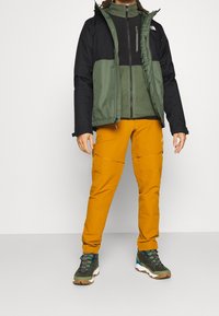 Chaqueta aislante negra y verde con cuello alto, pantalones naranja y botas de senderismo multicolores. Cuenta con bolsillos con cremallera y puños ajustables.