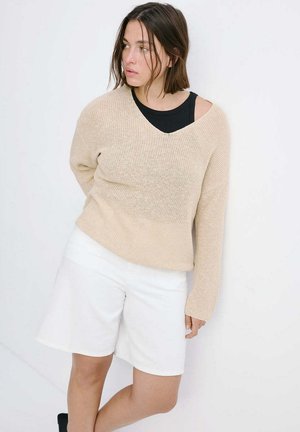 Kvinde med skulderlangt brunt hår læner sig mod en hvid væg iført en beige striktrøje over en sort top og hvide knælange shorts.