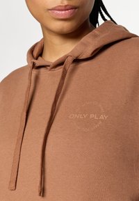 Brun hette med en myk tekstur, med en frontlomme i kengurustil og snor. "ONLY PLAY"-logoen er vist i et sirkulært design.