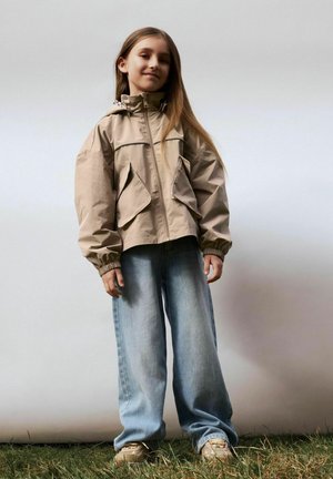 Jong meisje buiten staand, gekleed in een beige jas met capuchon, losse lichtblauwe spijkerbroek en beige sneakers, met lang stijl haar los gedragen.