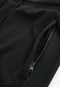 Gros plan d'un pantalon en tissu noir avec une poche zippée noire partiellement ouverte sur la couture latérale.