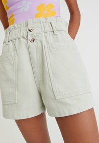 Světlé zelené džíny s vysokým pasem, dvěma předními kapsami a dvouknoflíkovým zapínáním. Tkanina má jemnou strukturu a strukturovaný design.