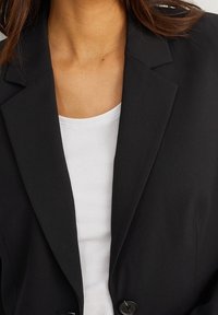 Femme portant un blazer noir sur une chemise blanche à col rond, montrant son clavicule et une partie de ses cheveux bruns.
